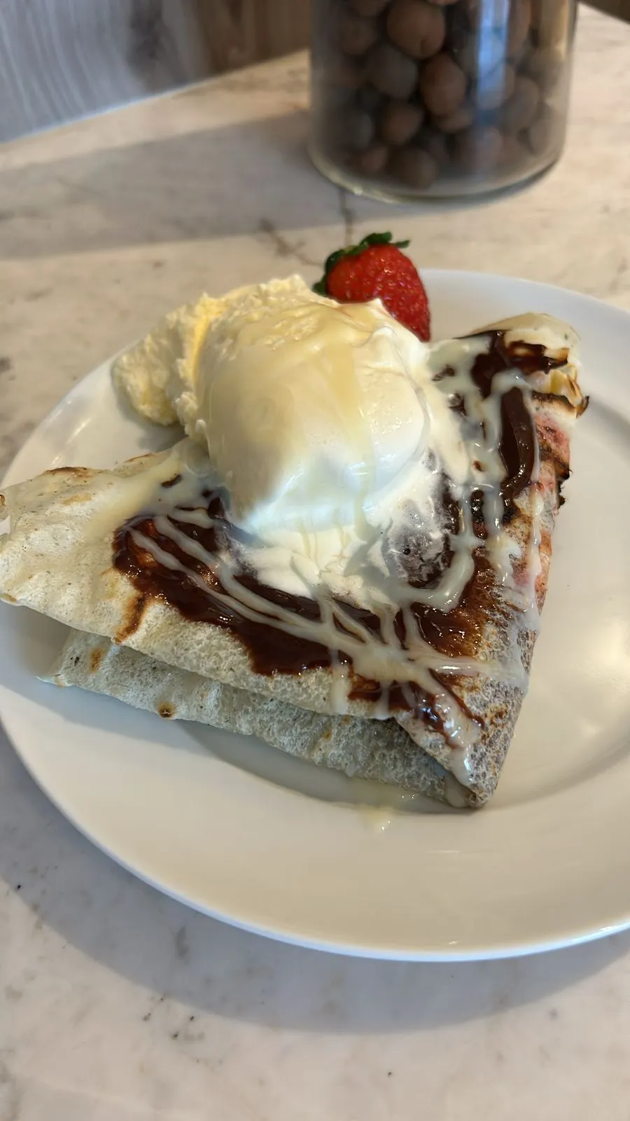 Plate with a folded crepe topped with chocolate and white sauce, a scoop of vanilla ice cream, a dollop of whipped cream, and a strawberry.