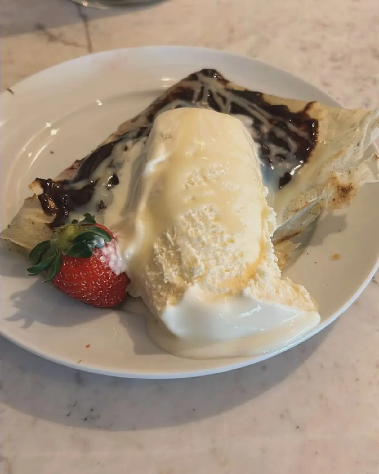 Plate with a folded crepe topped with melted vanilla ice cream and chocolate sauce, accompanied by a fresh strawberry.