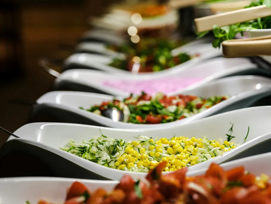 Close-up of a buffet line with white bowls filled with mixed salads including corn, greens, and tomatoes.