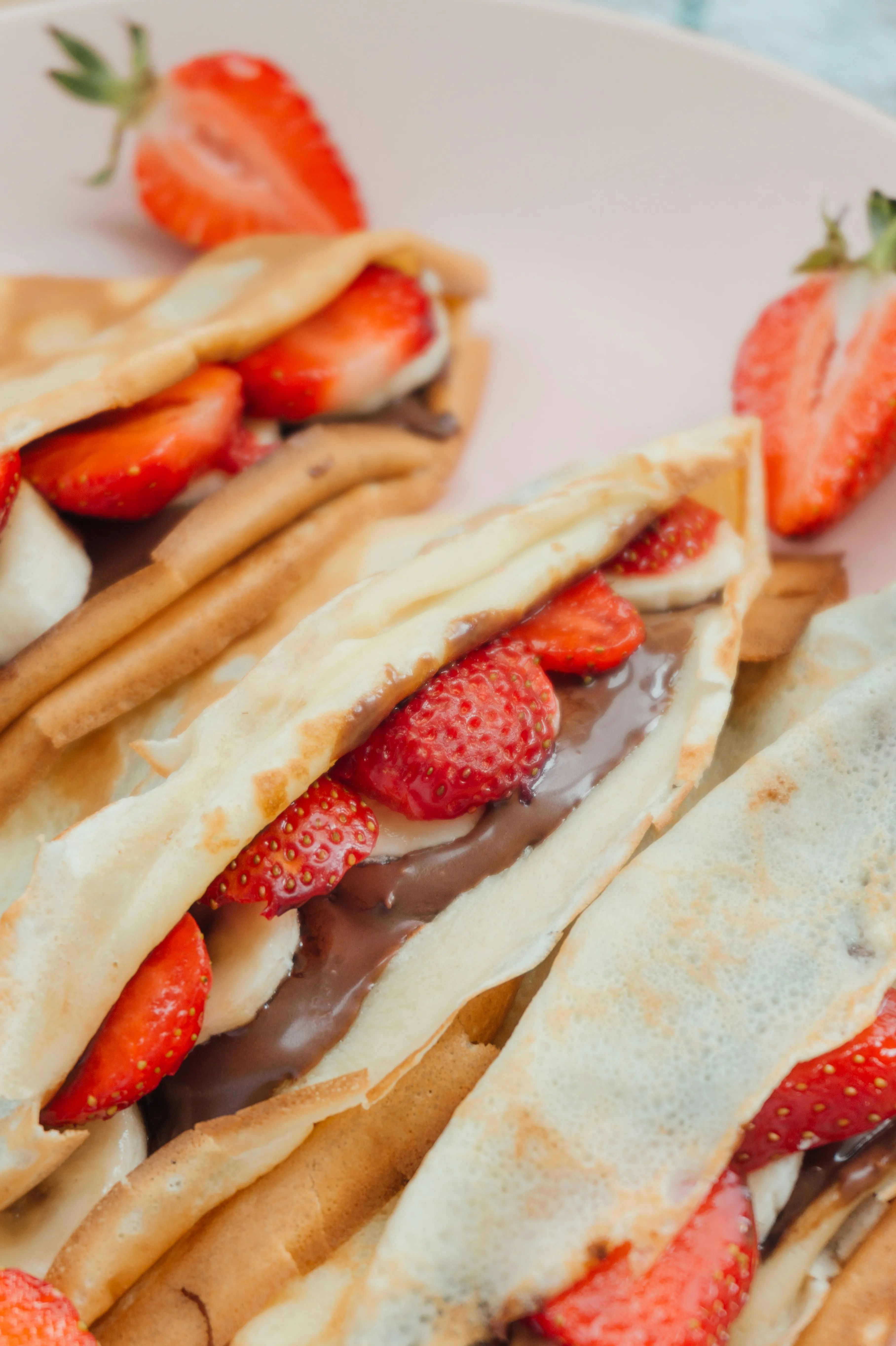 Close-up of crepes filled with sliced strawberries, banana, and chocolate spread on a pink plate.