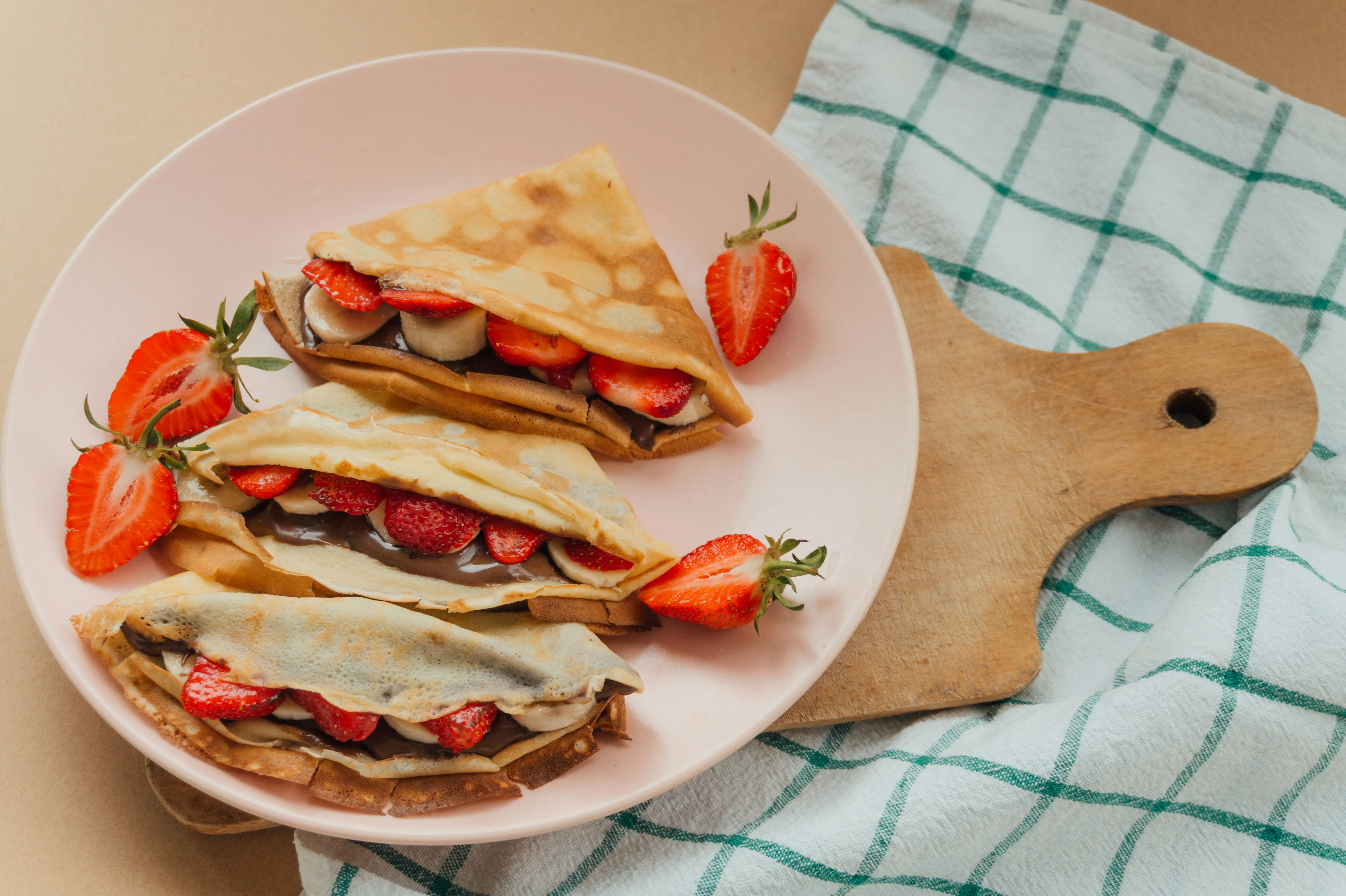 Three folded crepes filled with sliced bananas, strawberries, and chocolate spread on a pink plate, garnished with halved strawberries.