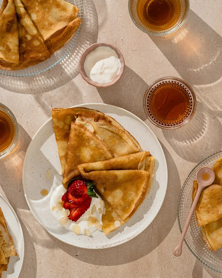 Plate with folded crepes topped with sliced strawberries, almond flakes, and cream, surrounded by glasses of tea on a beige surface.