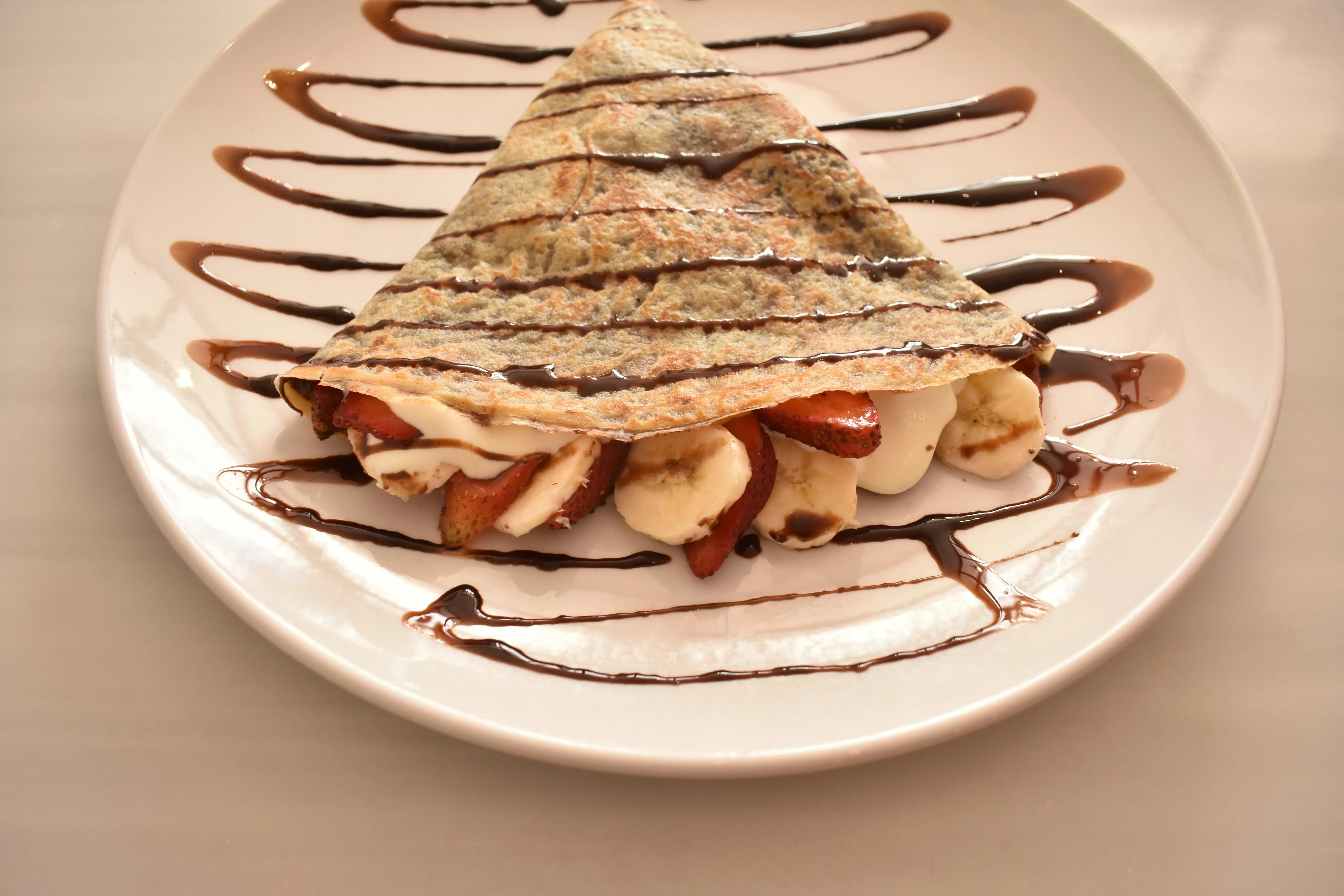 Crepe folded into a triangle filled with sliced strawberries, bananas, and cream, drizzled with chocolate sauce on a white plate.