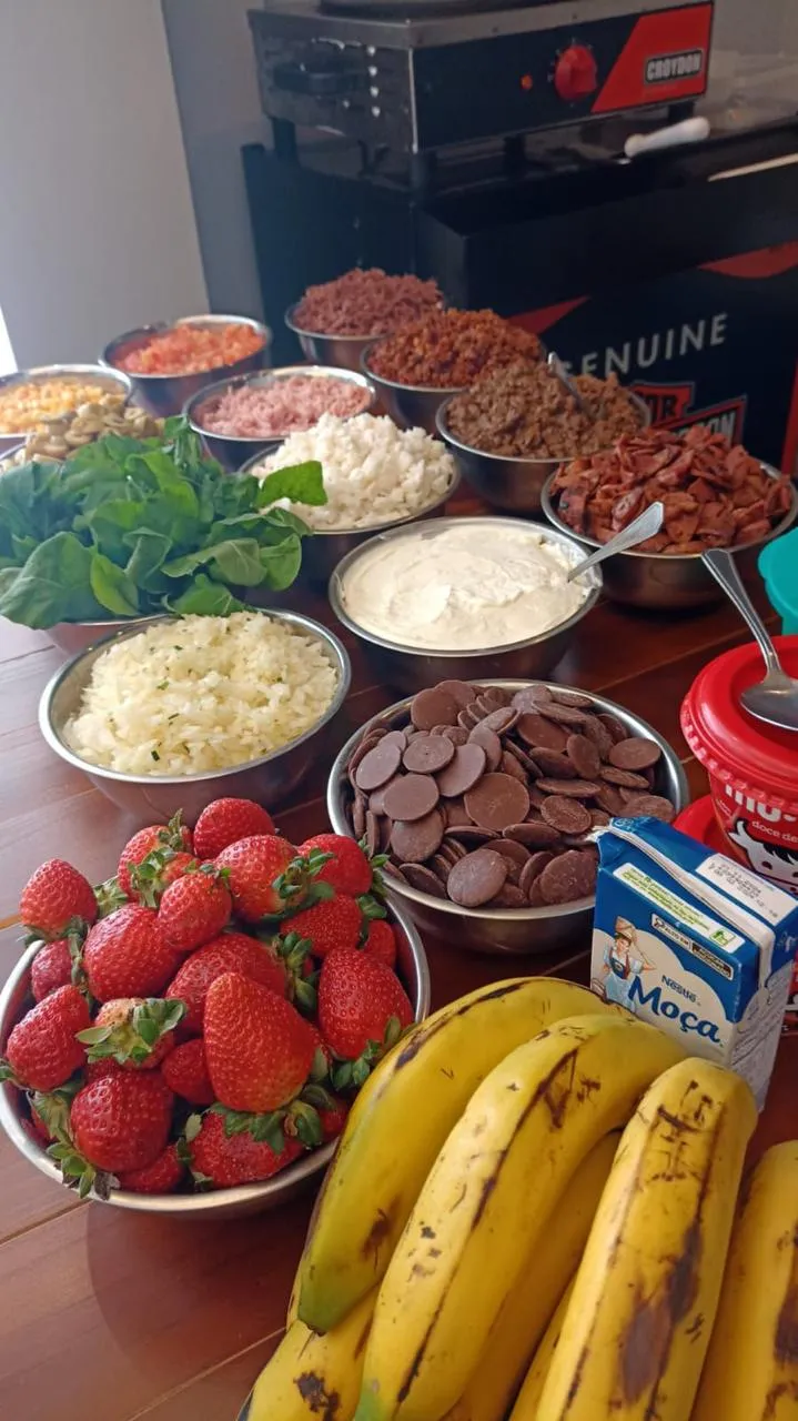 Bowls of fresh strawberries, bananas, leafy greens, shredded cheese, chocolate discs, cream, and various chopped meats and vegetables arranged on a wooden table.