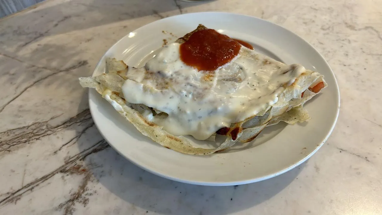 Savory folded crepe topped with white sauce and red salsa on a white plate over a marble table.