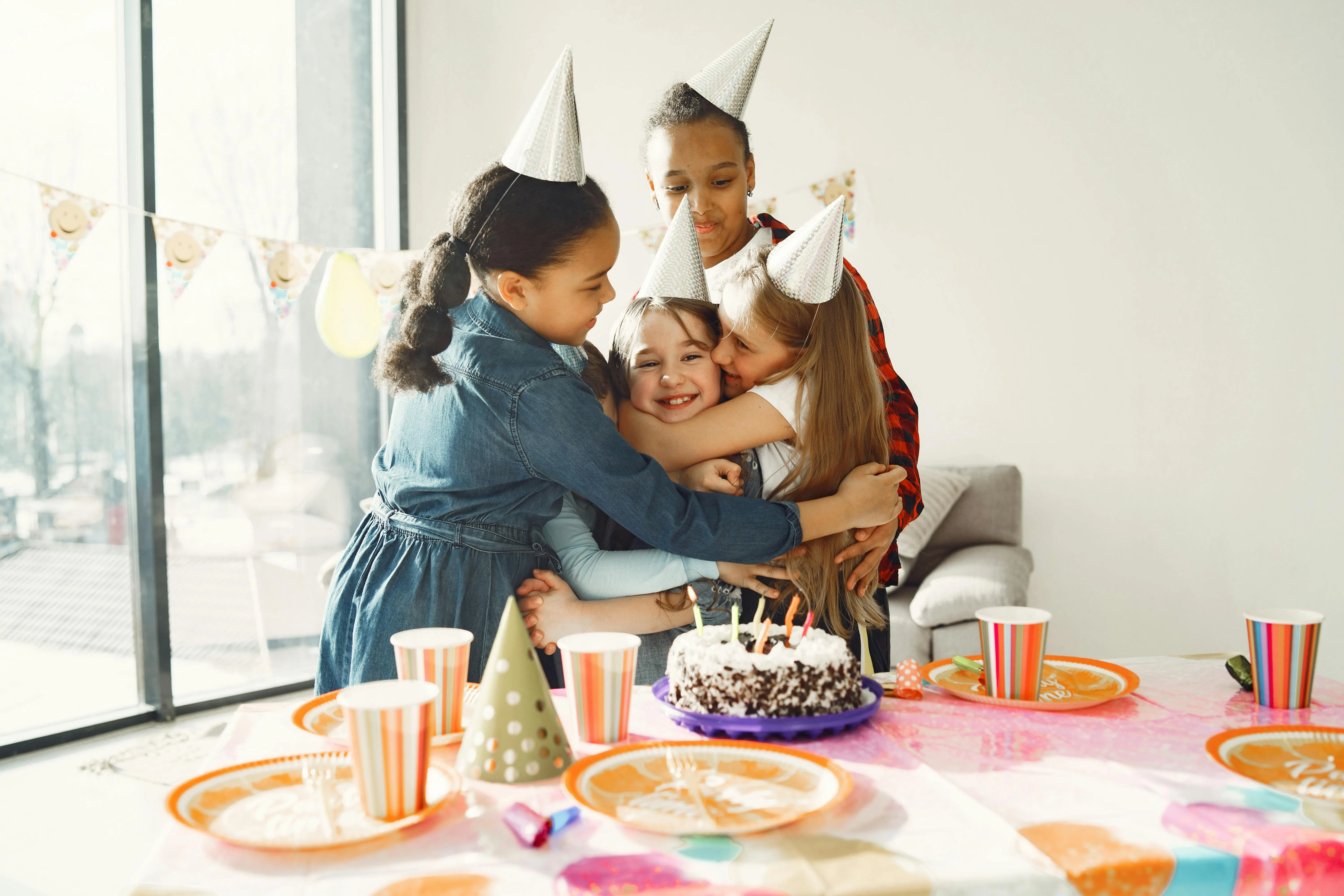 Four children wearing party hats hugging and smiling around a birthday cake on a decorated table.