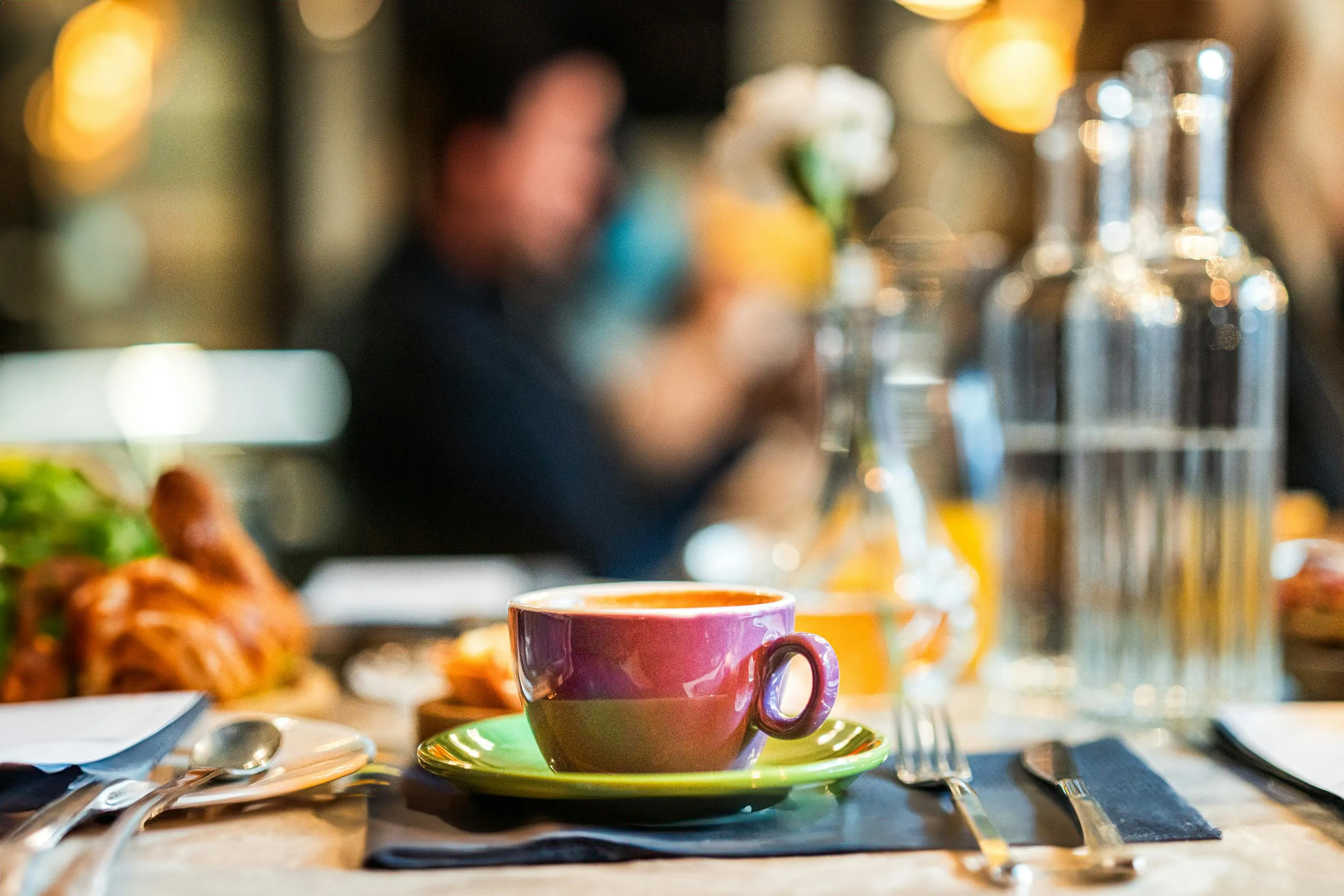 Purple coffee cup on a green saucer on a table set with utensils, with blurred people and glass bottles in the background.