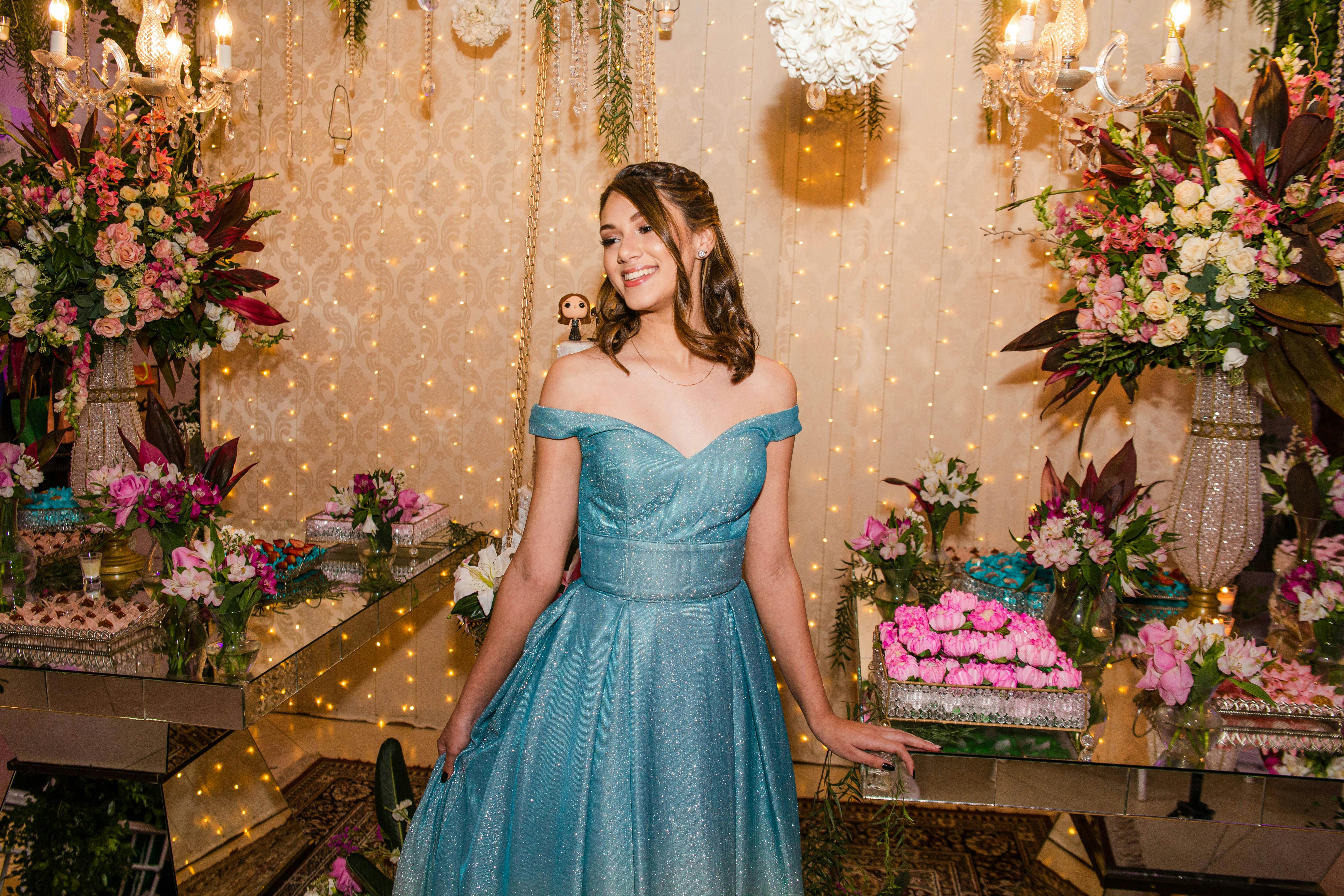 Young woman in an off-shoulder sparkling blue gown smiling in a decorated room with flowers and fairy lights.