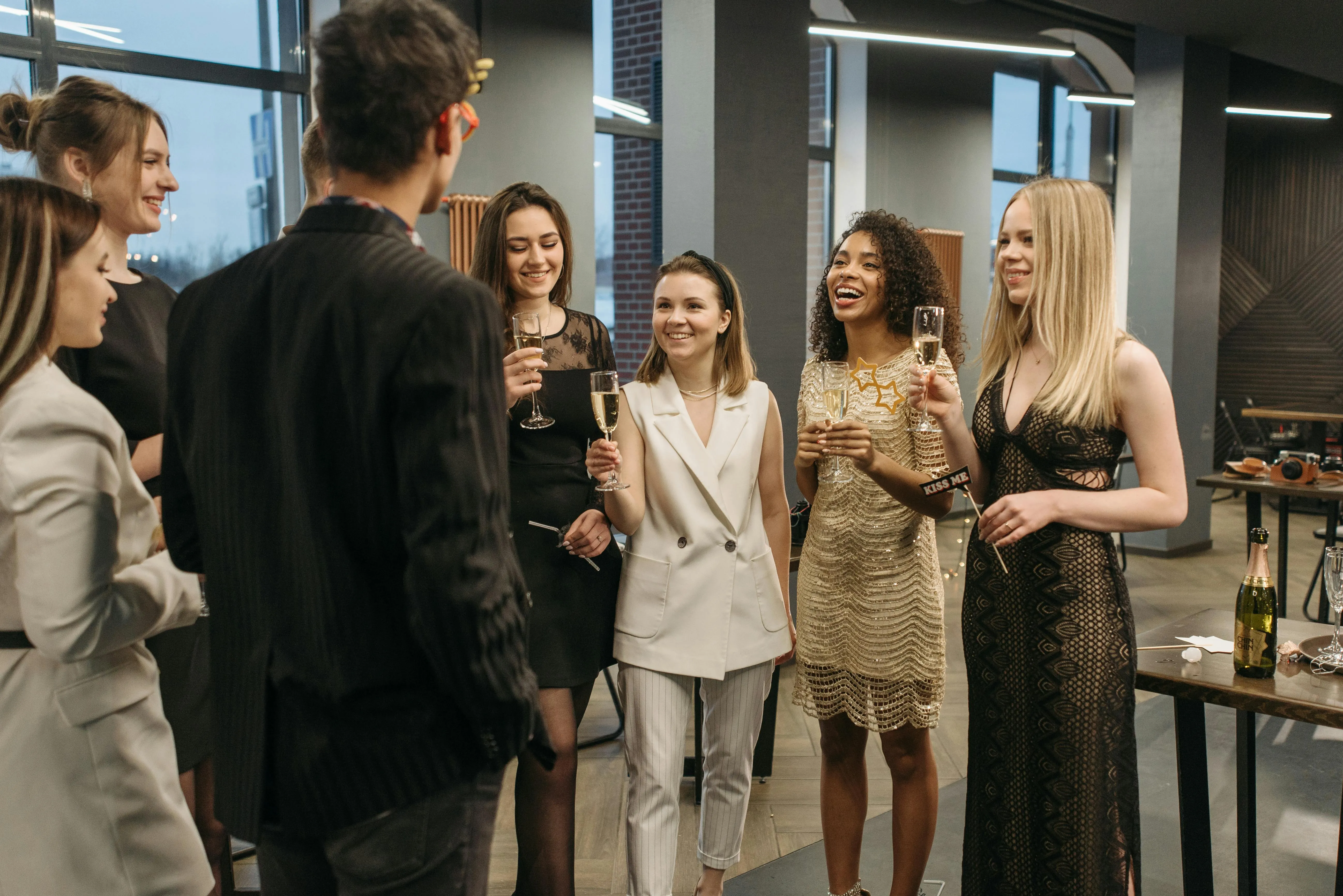 Group of six women and one man in stylish attire smiling and holding champagne glasses at an indoor party.