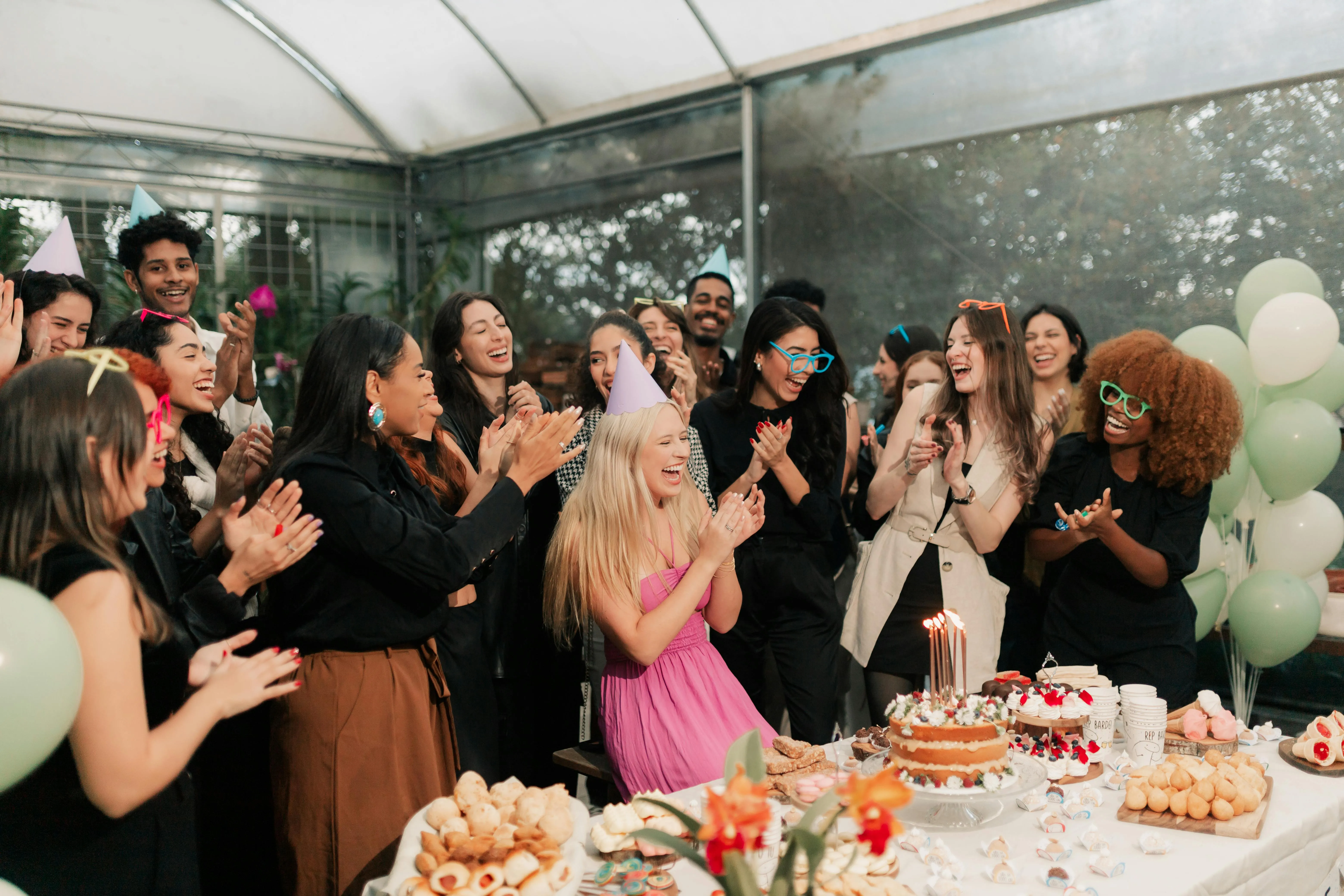 Group of diverse friends celebrating a birthday with a decorated cake and snacks, all clapping and smiling indoors.
