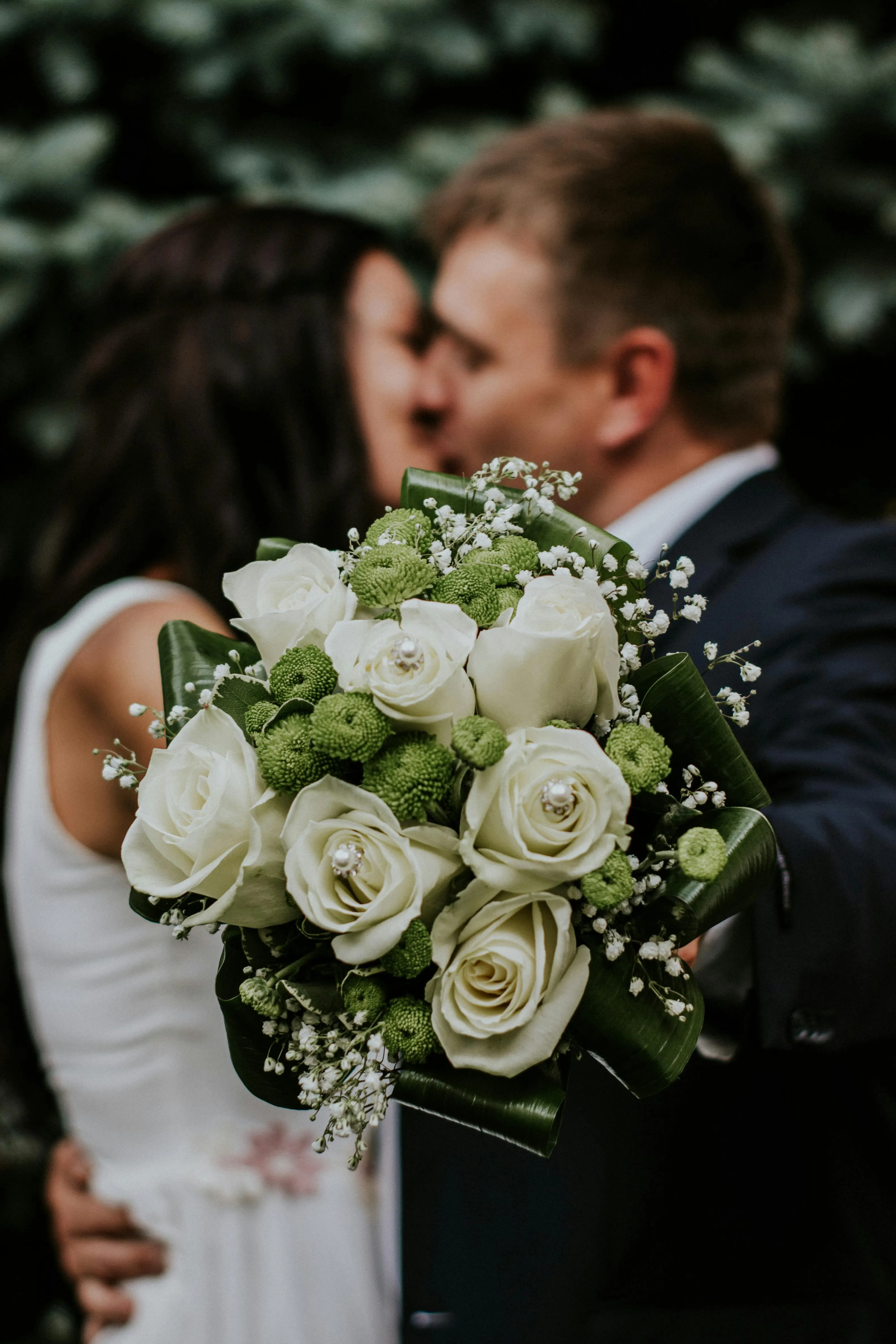 Close-up of a wedding bouquet with white roses and green accents held by a couple kissing in the background.